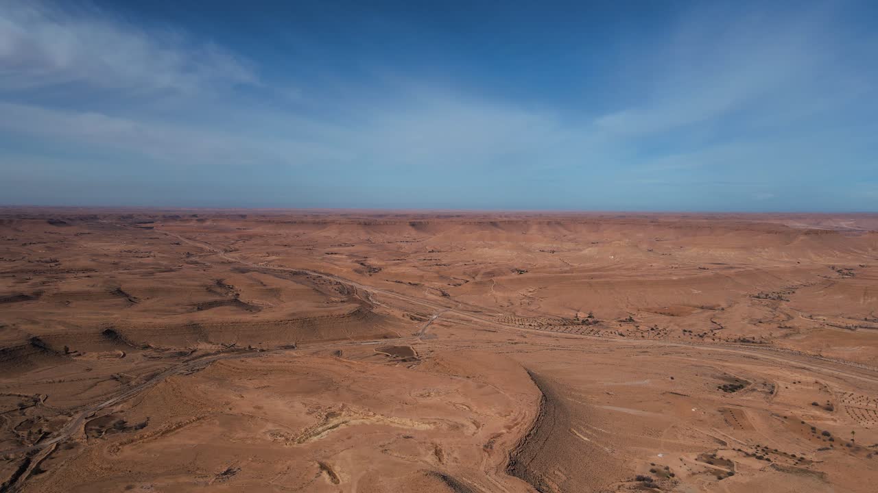 Expansive aerial view of the arid landscape near Ksar Guermessa, Tunisia. Red sand, sparse vegetation, and a vast horizon under a clear sky.