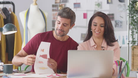 male and female fashion designers in studio making video call and showing designs at desk on laptop