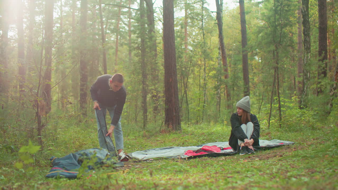 Young couple setting up for camping in forest, man squats to remove tent poles and toss pack aside while woman in beanie and jacket sits cross-legged on tent fabric watching him with warmth