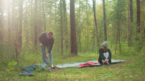 young couple setting up for camping in forest, man squats to remove tent poles and toss pack aside while woman in beanie and jacket sits cross-legged on tent fabric watching him with warmth