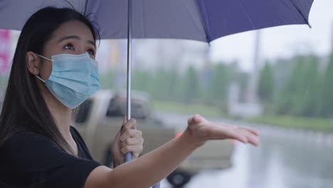 young asian woman wear surgical mask holding umbrella standing on the road side, on the raining day, shower rainy season tropical weather, reach out her hand feeling the rain drops, protect her self