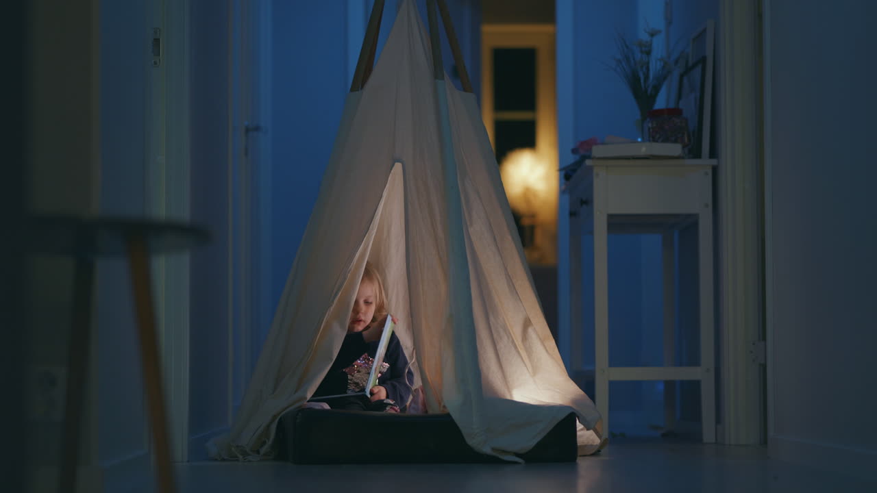 Video of a young intelligent girl reading book inside cozy indoor tent at night