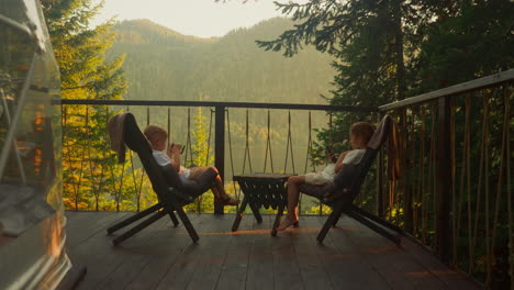 los compañeros se sientan en la mesa entreteniendo con teléfonos inteligentes. los niños en edad preescolar usan dispositivos sentados en el balcón en un clima soleado de verano. el video muestra el ambiente para relajarse