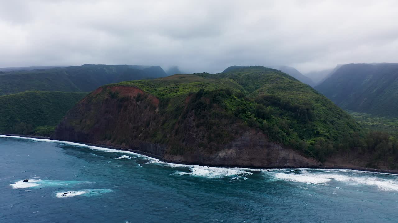 Sweeping green cliffs curve along a rugged coastline as deep blue waves break below, framed by thick gray clouds drifting overhead in this striking wild landscape filled with natural drama.