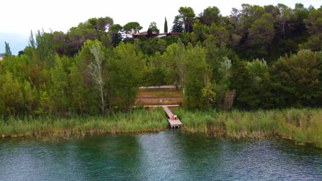 Volando-Lejos-De-Una-Pareja-Sentada-En-Una-Plataforma-De-Madera-Frente-A-Un-Lago-Con-Agua-Clara,-Entre-Los-árboles-Y-La-Casa-En-El-Fondo,-En-Banyoles,-Cataluña,-España