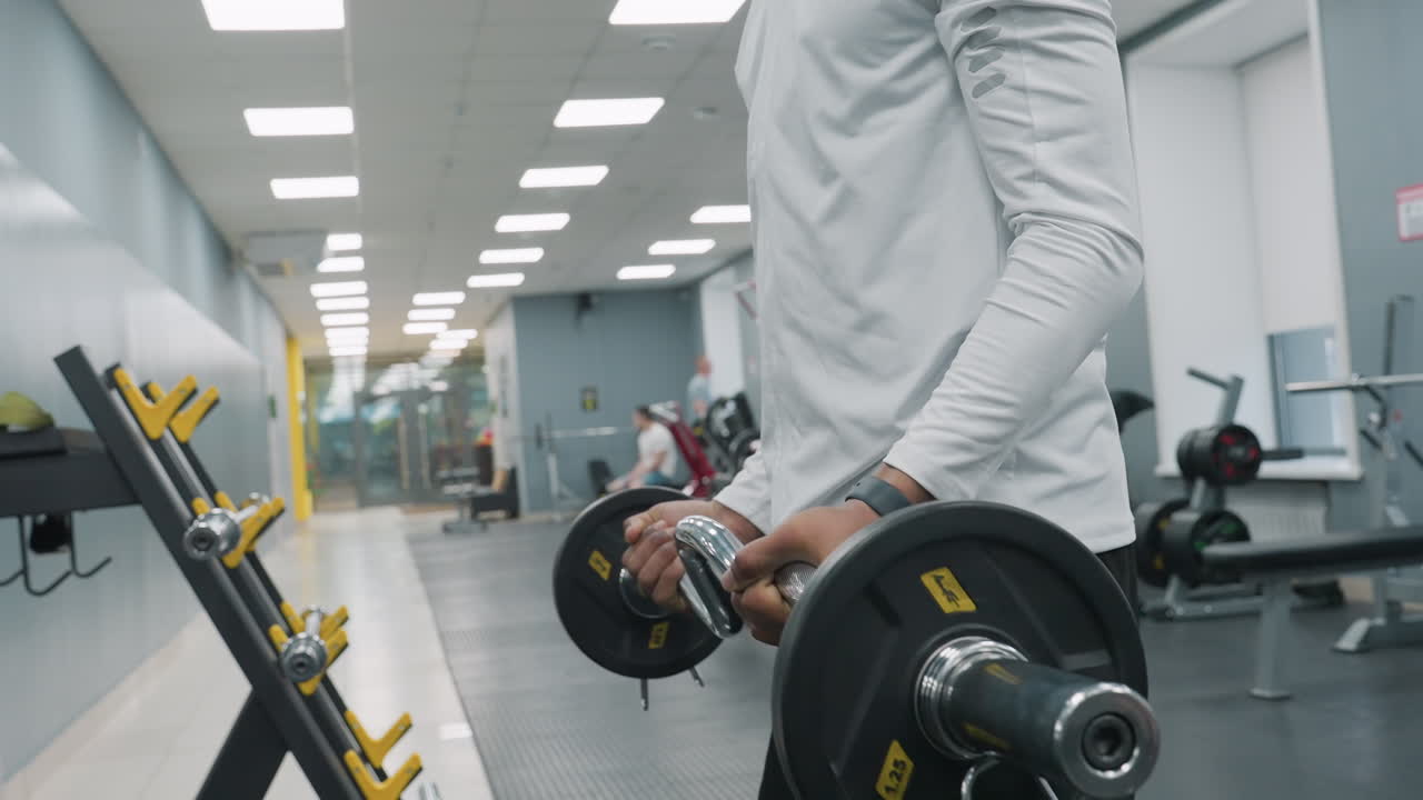 Side view of man lifting barbell loaded with weight plates while other gym members work out in background, showing hands gripping chrome bar and focused strength training session on rubber flooring