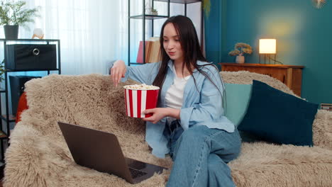 woman happily eating popcorn watching movie on laptop at home sofa emotionally laughing enjoyment