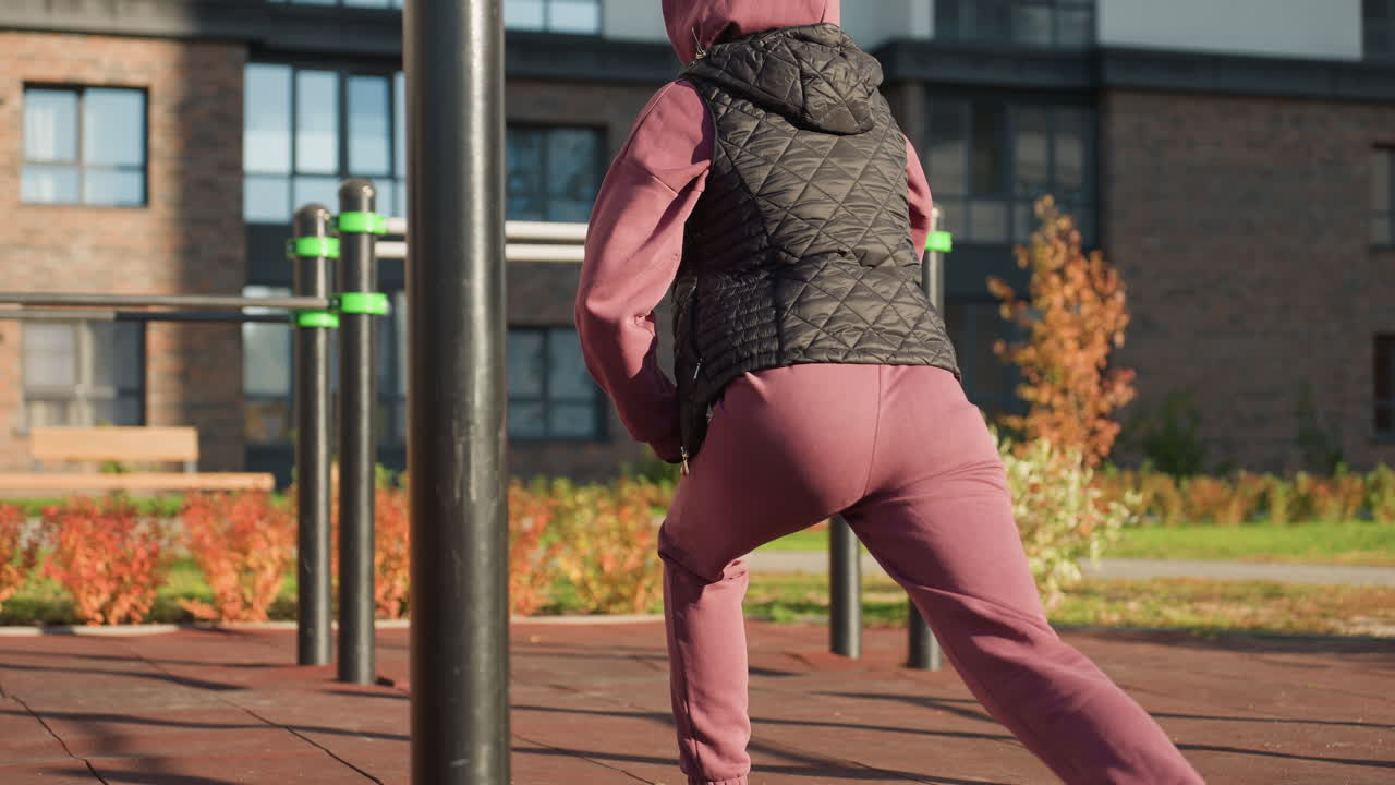 Back view of active woman in sports wear doing split squats with hands placed on legs in modern urban park environment featuring flowers rails shadows on rubber tile under bright sunlight