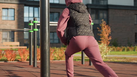 back view of active woman in sports wear doing split squats with hands placed on legs in modern urban park environment featuring flowers rails shadows on rubber tile under bright sunlight