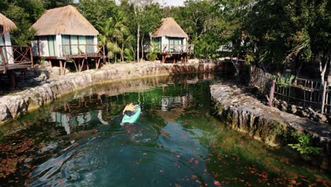 Aerial-view-over-a-person-kayaking-on-a-river-flowing-through-luxury-beach-wooden-houses-in-a-tropical-resort-surrounded-by-trees,-in-Tulum,-Mexico