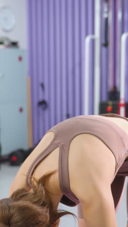 joven con ropa deportiva marrón practicando de pie hacia adelante inclinándose en el gimnasio su cabello cae suavemente sobre los hombros, mejorando su comportamiento enfocado y pacífico en medio de equipos de gimnasio modernos