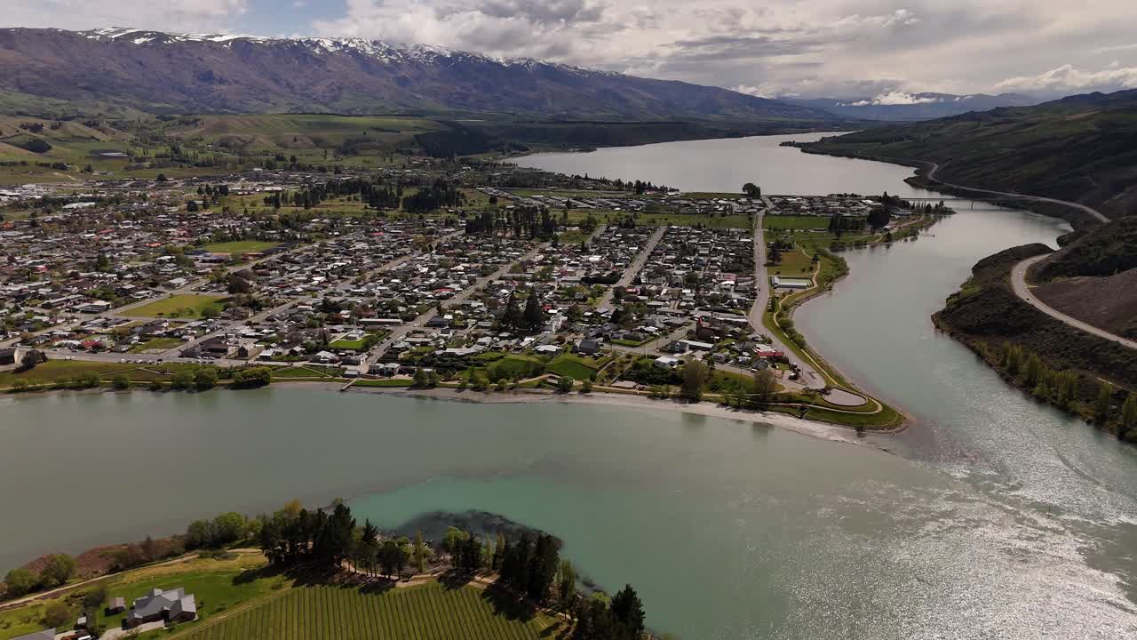 Cromwell Town in Otago Region facing Clutha River with turquoise water curves around bright settlement. Rugged South Island mountains under soft daylight in autumn season. Aerial wide shot