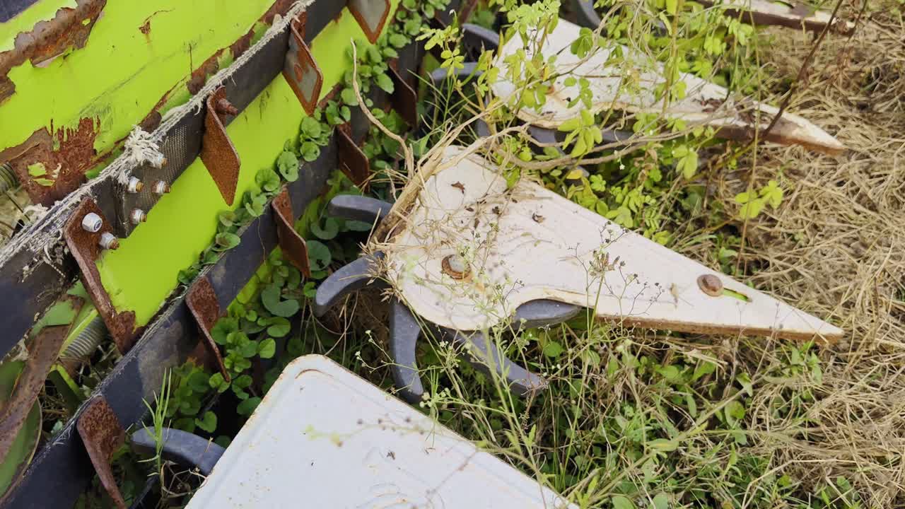 A tilt-up shot revealing the rusted metal blades and gears of an old harvester machine, partially covered in dry grass and weeds, symbolizing the passage of time in rural agricultural landscapes