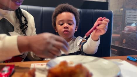two year old black baby, mix raced, learns how to use the fork eating chicken in a restaurant seated next to his mother
