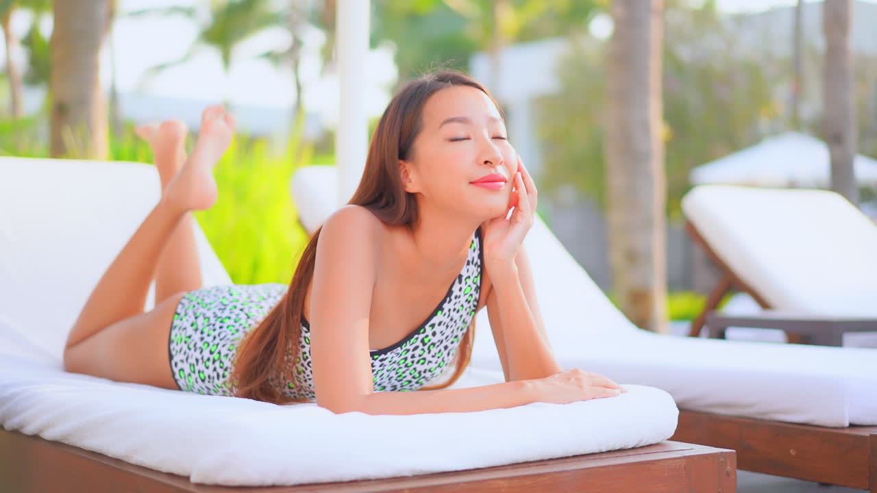 화려한 열대 리조트 미소, 주간 슬로우 모션에서 편안한 데크 의자에 그녀의 배에 누워 얼룩덜룩 한 모노 키니에 젊은 아시아 소녀