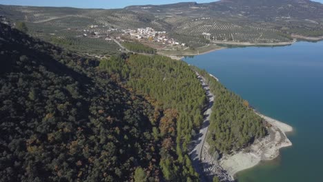 Aerial-view-of-a-road-surrounded-by-a-forest-close-to-a-lake-with-a-small-village-in-the-horizon