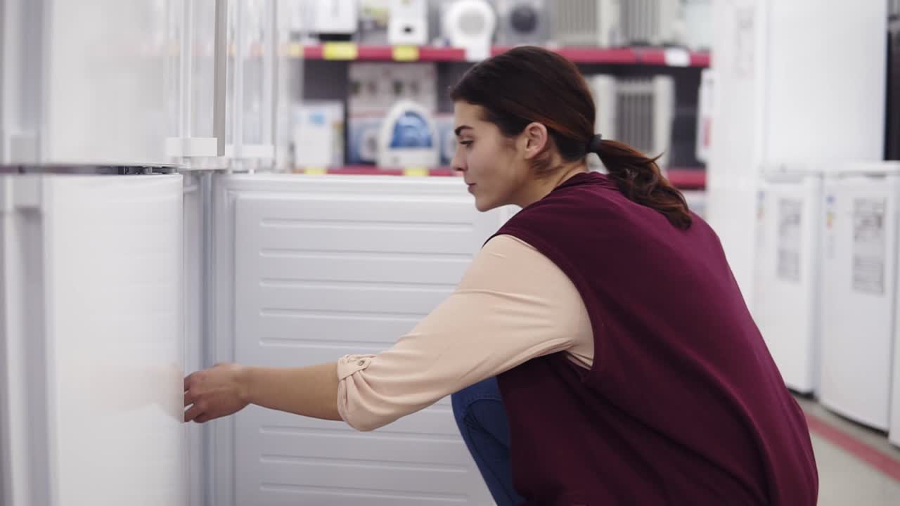 Young girl in her 20's comes up to a fridge in hardware store opens the freezer and checks out what's inside. Looking for household equipment