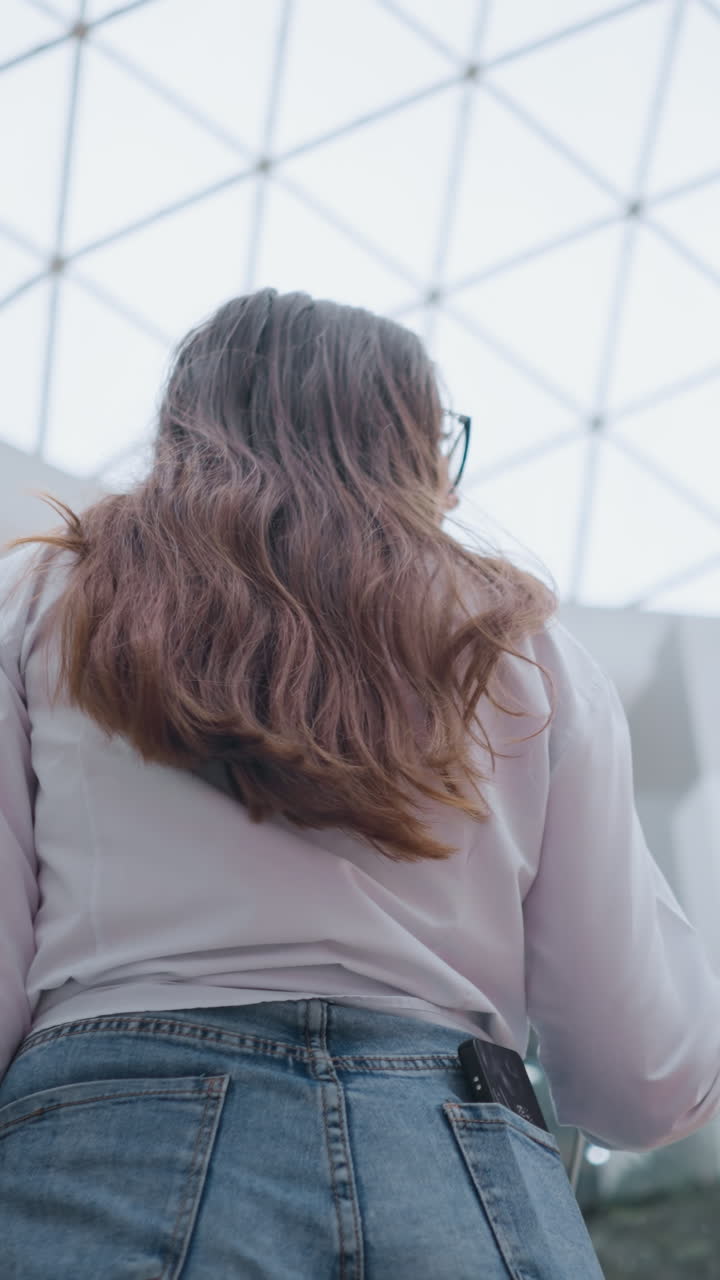 현대 쇼핑몰에서 이동하는 에스컬레이터에서 젊은 여자의 뒷면, 뒷 주머니에 전화, 레일에 손, 다른 사람들이 앞에 올라, 일상적인 쇼핑 경험과 도시 생활 방식을 보여줍니다.
