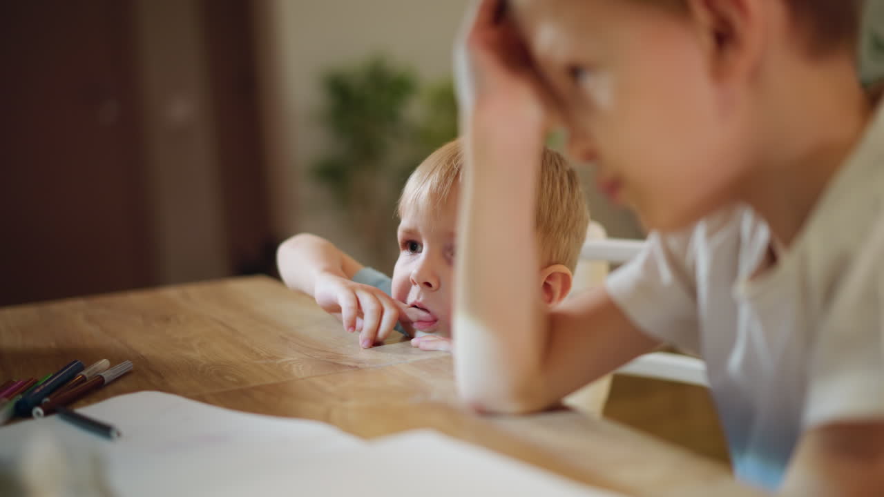 Tired little boy rests head on wooden table near scattered colorful markers and open notebook while older sibling points towards something with hand gesture, scene of childhood moment inside cozy home