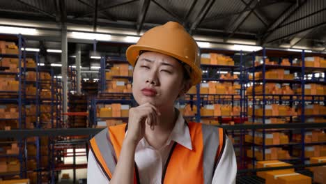 close up of asian female engineer with safety helmet standing in the warehouse with shelves full of delivery goods. thinking about something and looking around in the storage