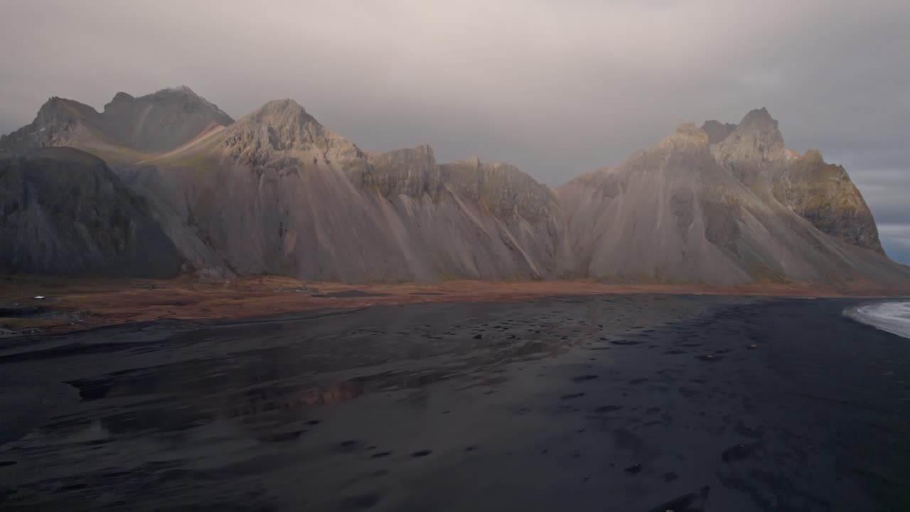 An evocative aerial shot captures the iconic Vestrahorn mountains rising dramatically behind the vast, volcanic black sand beach of Stokksnes on Iceland's southeastern coast