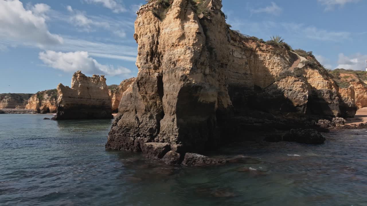 Aerial drone performs a helix orbit around dramatic limestone sea arches and layered cliffs, with water sparkling in the sun near Lagos, Algarve, Portugal
