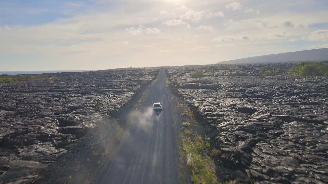 Aerial tracking of a car navigating a dirt road amid a lava field on Hawaii's Big Island, with a smoky sunset forming the backdrop