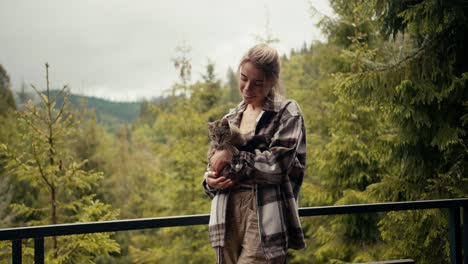 Happy-blonde-girl-petting-her-cat-on-the-balcony-of-a-country-house-overlooking-a-coniferous-forest-in-the-mountains