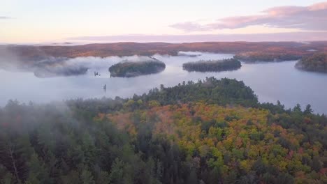 plano general del amanecer aéreo volando sobre los colores del bosque otoñal en la cresta rocosa brumosa se desplaza hacia el lago brumoso con islas de pinos cubiertas de niebla y nubes rosadas en kawarthas ontario canadá