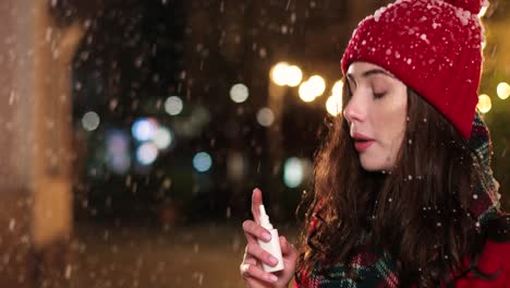 vista de cerca de una mujer caucásica enferma con abrigo rojo al aire libre usando spray nasal en la calle mientras nieva en navidad