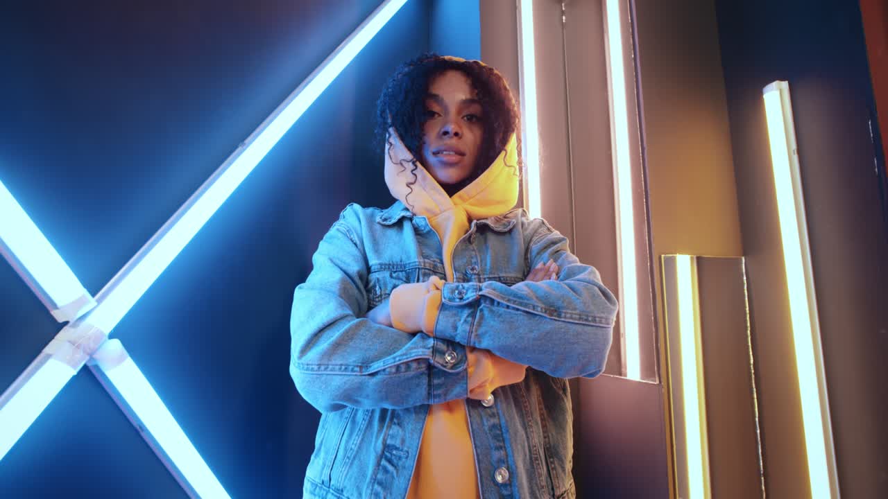 Wide angle portrait of stylish urban african american black woman in yellow hoodie and jeans jacket standing by wall in night disco club backlit with purple and blue neon light stripes. Urban style