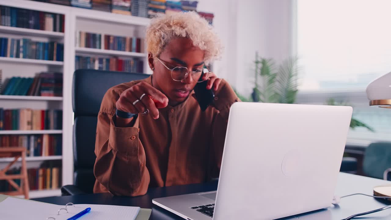 Afrikansk kvindelig medarbejder med briller arbejder på kontor og taler i mobiltelefon.