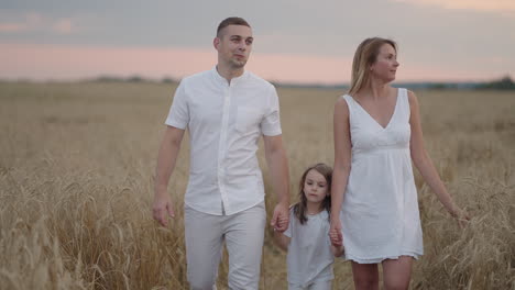 young couple of parents with girl children holding hands of each other and running through wheat field at sunset. happy family jogging among barley meadow and enjoying nature together. slow motion