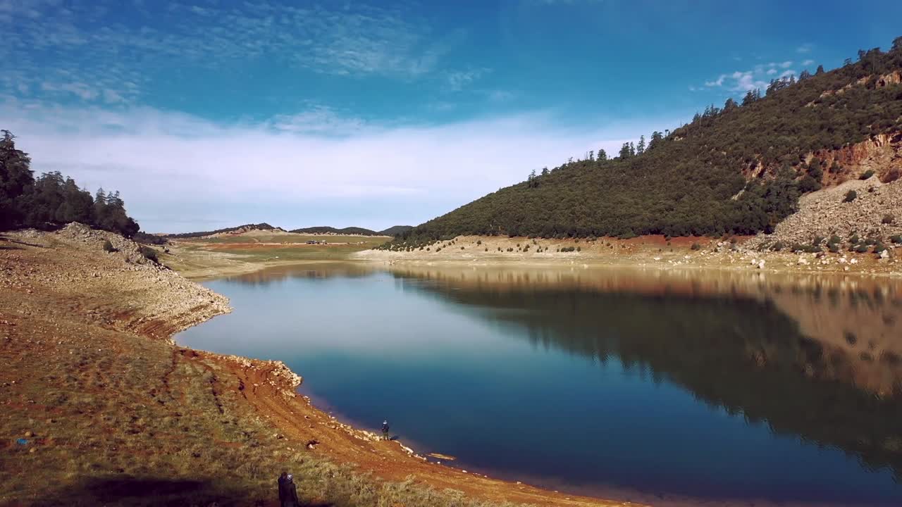 Ampia veduta aerea di un sereno lago di montagna incastonato tra le colline. Presenta acque blu che contrastano con le rive rossastre e la vegetazione verde nel Medio Atlante.