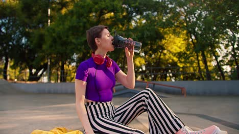 a girl with a short haircut with a purple top and striped pants in pink roller skates sits in a skate park on a concrete floor and drinks water from a special bottle