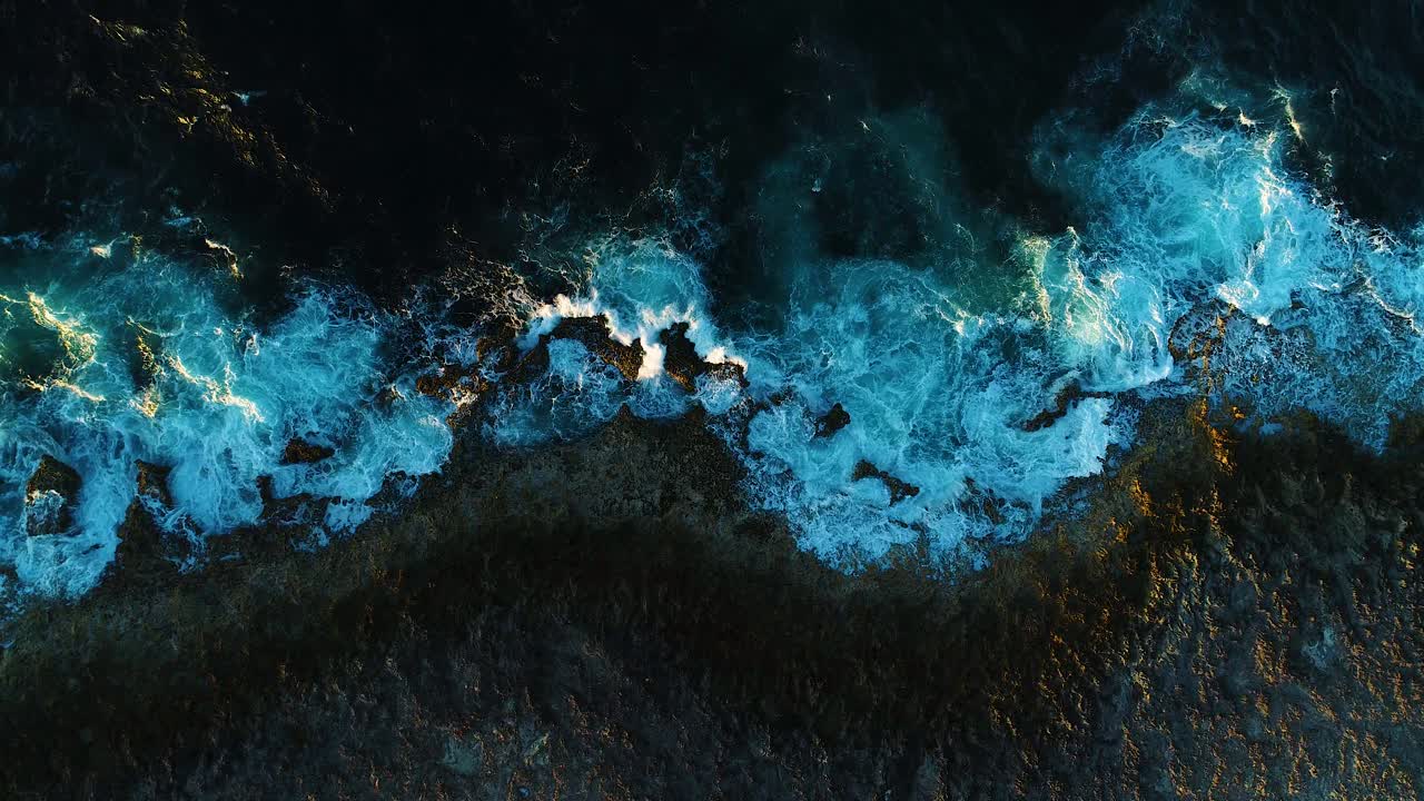 Static rocky volcanic ocean shoreline, golden hour light glows on wave ripples aerial drone