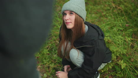 young woman in casual outdoor outfit squats on grassy forest ground while smiling warmly and gazing affectionately at someone standing in front of her during peaceful moment in nature
