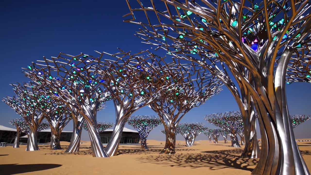 Metallic sculptural trees featuring vibrant glass foliage rising from sandy desert terrain, reflecting innovative design against cloudless Dubai skyline