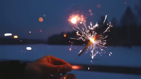 snowy winter landscape glowing at dusk, hand holding burning sparkler with colorful fireworks exploding against dark twilight sky and distant forest backdrop