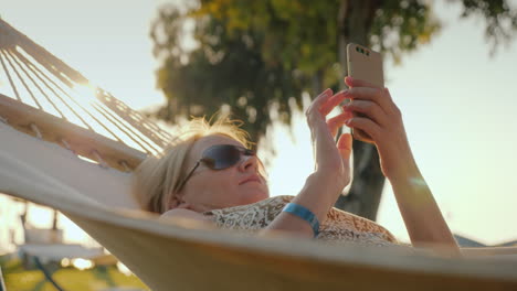 una mujer usa un teléfono inteligente de vacaciones y se relaja en una hamaca en el video de 4k temprano en la mañana