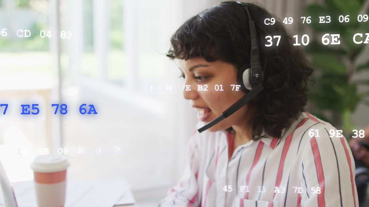Woman on headset taking tech call, leaning and typing while hex code overlaying laptop and papers