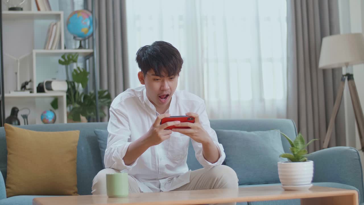 Asian Man Celebrating Winning Game On Smartphone While Sitting On Sofa In The Living Room
