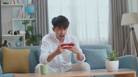 asian man celebrating winning game on smartphone while sitting on sofa in the living room