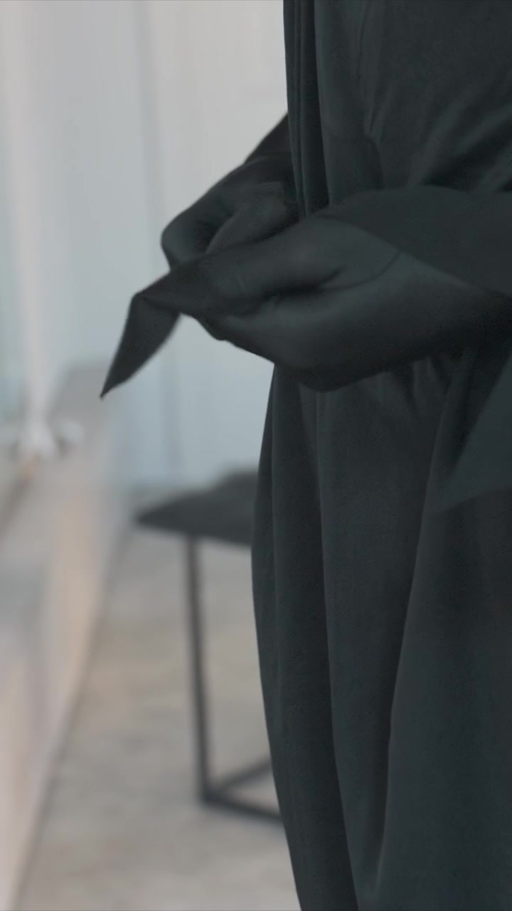 Primo piano di una persona con guanti neri che maneggia del materiale. Le mani muovono il materiale avanti e indietro, suggerendo un'azione di legatura. Un ambiente interno semplice con pareti e pavimento di colore chiaro è visibile per tutta la scena.