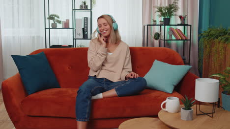 happy young woman in wireless headphones relaxing sitting on sofa at home listening favorite music