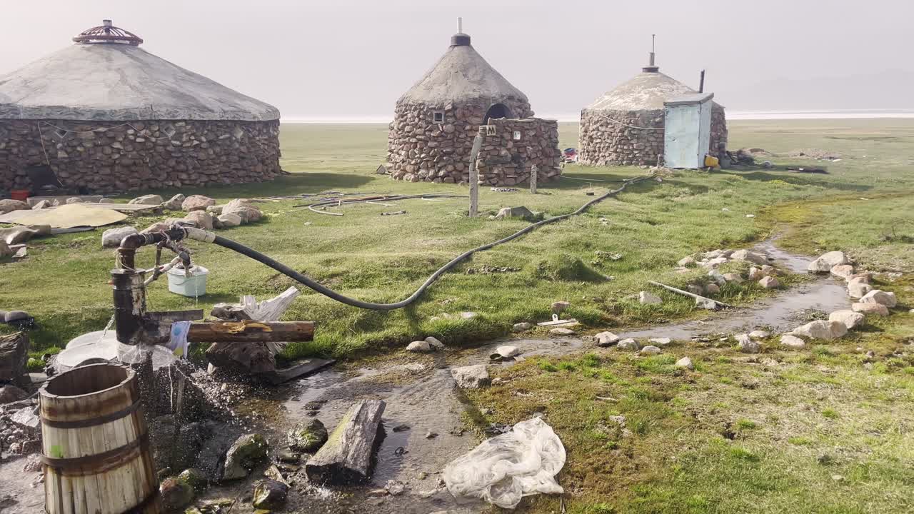 Iurte tradizionali in pietra e una pompa per pozzo d'acqua nell'isolato altopiano del Kirghizistan, che mostrano l'architettura nomade rustica e la vita rurale vicino al lago Song-Kul in Asia Centrale
