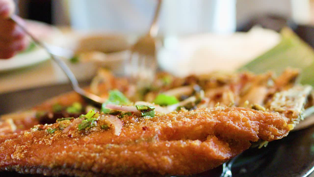 Close-up of plated crispy fried fish garnished with herbs, fork approaching, natural restaurant lighting