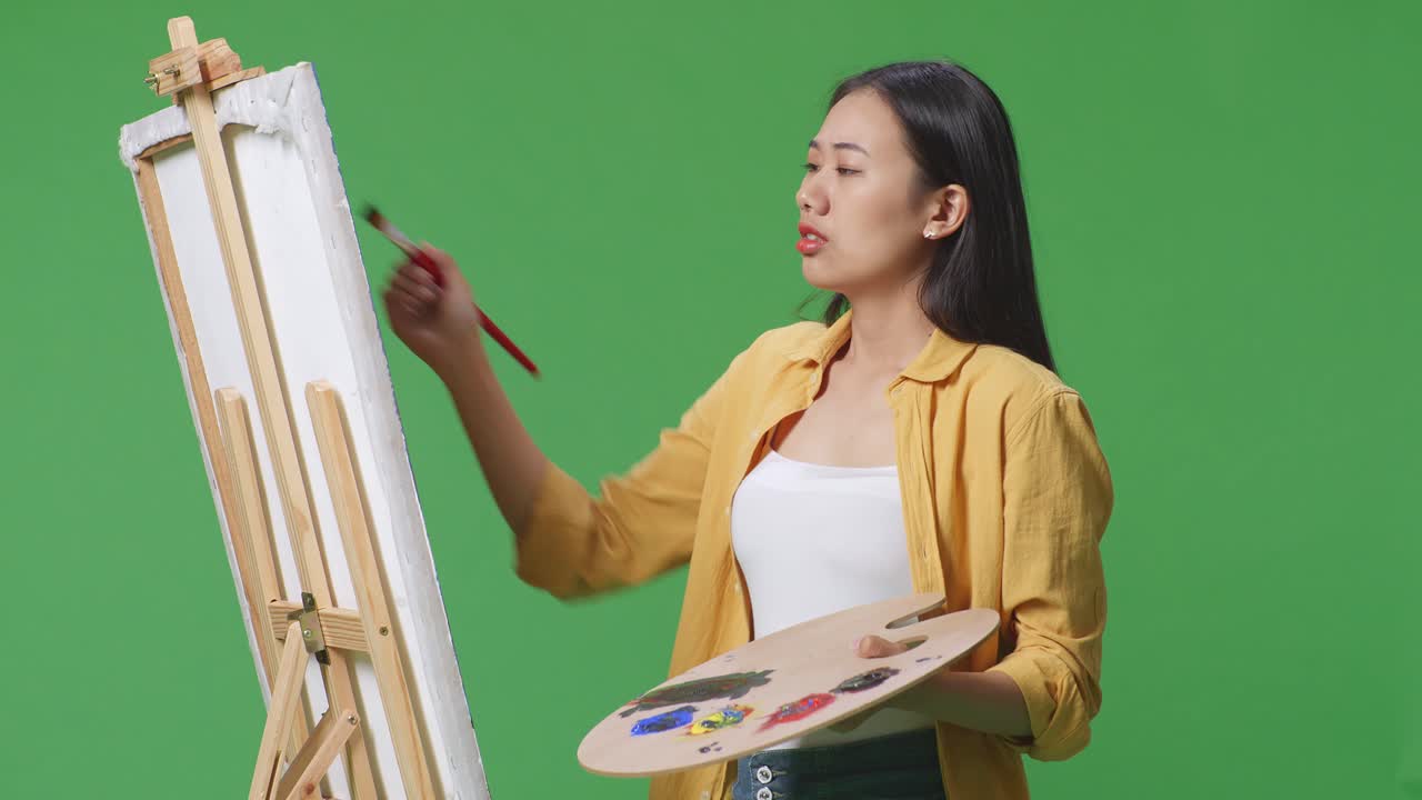 아시아 여성 예술가가가 머리를 흔들고 머리가 아파서 녹색 스크린 스튜디오에서 오일 페인트와 으로 캔버스에 그림을 그리고 있습니다.