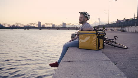 happy food delivery guy rests at sunset next to his thermal backpack looking at the beautiful scenery on the shore of a river