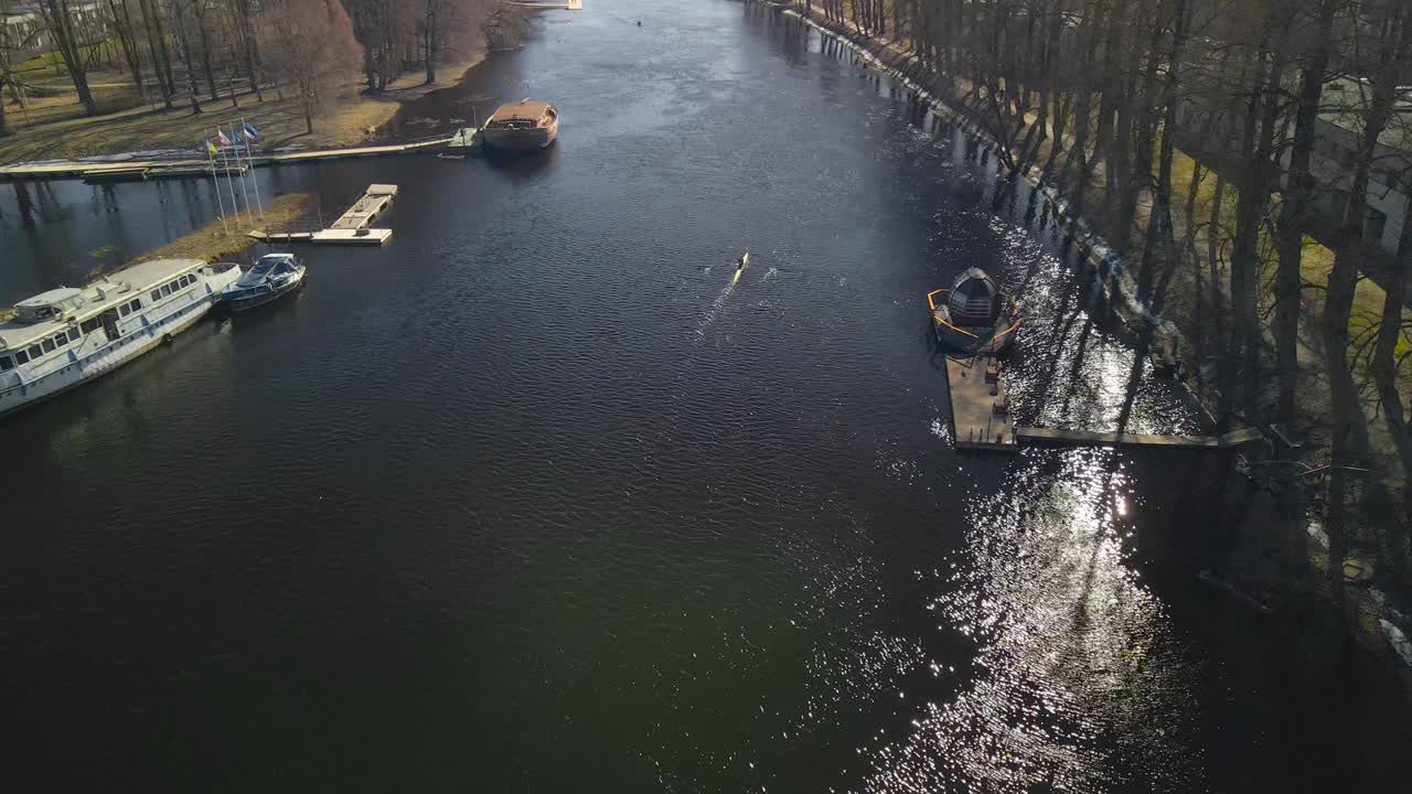 Aerial drone footage flying lower on top of Tartu cold dark colored river called Emajõgi where people are rowing a kajak or a paddle boat during spring time sunny day and large boats are achored too.